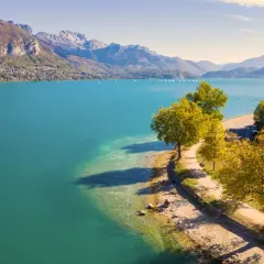 Vue aérienne du lac d'Annecy, ses eaux turquoise bordées par une route sinueuse et des arbres aux couleurs d'automne. La taille du lac est impressionnante.