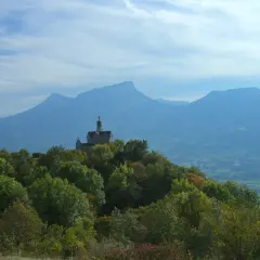 Un petit Mont Saint Michel savoyard, une chapelle perchée sur une colline boisée, domine un paysage de montagnes bleutées sous un ciel nuageux.