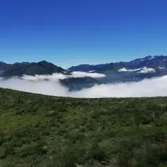 Vue panoramique depuis le col de Riou, avec des montagnes verdoyantes émergeant d'une mer de nuages sous un ciel bleu éclatant.