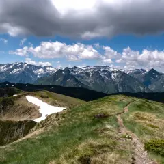Sentier rocailleux menant vers le pic de Tarbesou, avec des montagnes enneigées en arrière-plan sous un ciel nuageux.