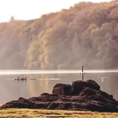 Épée plantée dans un rocher au bord du lac de Trémelin, entouré de brume et de collines boisées.