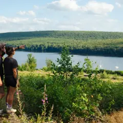 Couple randonneurs admirant le lac des Vieilles Forges, avec des voiliers sur l'eau et une forêt dense en arrière-plan.