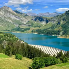 Vue panoramique du lac de Roselend, un barrage imposant bordé de forêts verdoyantes et de montagnes majestueuses sous un ciel bleu éclatant.