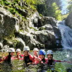Groupe souriant en combinaison néoprène, casques blancs et bleus, profitant d'une descente en canyoning près d'une cascade. Une belle aventure de canyoning dans le Vallon Pont d'Arc.
