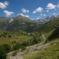 Vue panoramique d'un col de tentes dans les Pyrénées, avec une route sinueuse traversant des prairies verdoyantes et des montagnes majestueuses sous un ciel bleu parsemé de nuages.