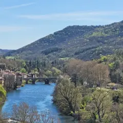 Vue panoramique de la gorge de l'Aveyron avec un village pittoresque, une rivière bleue et des collines verdoyantes sous un ciel clair.