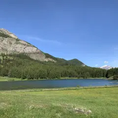 Le lac de Barbeyroux, paisible, reflète le ciel bleu. Des montagnes rocheuses et une forêt dense bordent ses rives verdoyantes.