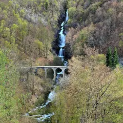 La cascade du Bief de la Ruine dévale une falaise rocheuse, traversée par un pont en arc. La nature verdoyante environnante annonce le printemps.