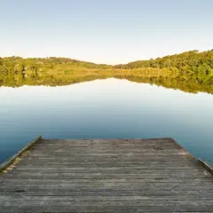Un ponton en bois mène au lac Marion de Biarritz, reflétant les collines verdoyantes sous un ciel clair.