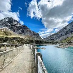 Vue panoramique du lac des gloriettes, un barrage imposant et des montagnes majestueuses sous un ciel bleu et nuageux.