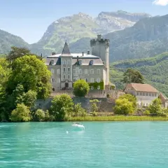Un château majestueux surplombe un lac turquoise, invitant à aller à Annecy. Des montagnes verdoyantes en arrière-plan.