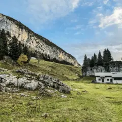 Un chalet blanc se dresse dans un pré verdoyant au pied d'une falaise imposante. Le col de l'Alpette offre un paysage alpin paisible.