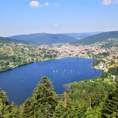 Vue panoramique du lac de Gérardmer, avec des voiliers sur l'eau bleue, entouré de collines verdoyantes et d'une ville pittoresque.