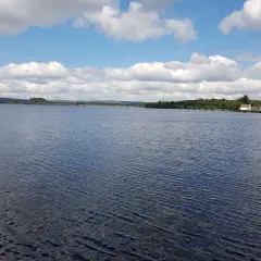 Vaste étendue d'eau calme sous un ciel bleu parsemé de nuages blancs. Le lac de Brennilis s'étend à perte de vue, bordé par une ligne d'arbres verdoyants et quelques constructions.
