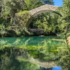 Pont de pierre envahi par la végétation au-dessus des eaux turquoise des gorges de la Siagne, reflétant le ciel et la végétation luxuriante.