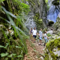 Une famille explore les **gorges des Tines**, un sentier étroit bordé de falaises rocheuses couvertes de mousse et de végétation luxuriante.