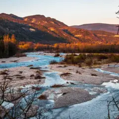 Le **saut de la Drôme** offre un paysage de rivière aux eaux bleues vives serpentant entre des bancs de galets, sous des montagnes dorées par le soleil couchant.