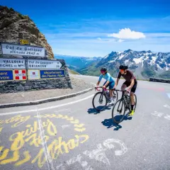Deux cyclistes gravissent le col du Galibier, un paysage alpin spectaculaire sous un ciel bleu éclatant.