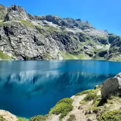 Le lac de Caillauas, d'un bleu profond, reflète les montagnes escarpées et verdoyantes sous un ciel clair. Une structure rocheuse rappelle un château sur la rive.