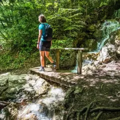 Une randonneuse traverse un pont en bois près de la cascade de Morette, entourée d'une végétation luxuriante.