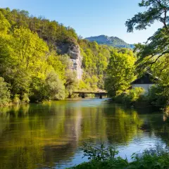 Rivière paisible reflétant la verdure luxuriante, un pont discret et une maisonnette au bord de l'eau, le tout sous un ciel bleu clair, évoquant la sérénité de la cascade de Syratus.