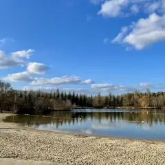 Plage de sable fin au bord d'un lac paisible, avec des familles profitant du soleil. La **cascade** de la **forêt de Fontainebleau** se reflète dans le **lac** sous un ciel bleu parsemé de nuages.