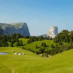Un campement de tentes et de randonneurs sur une prairie verdoyante, avec des montagnes imposantes en arrière-plan. Le **pas de l'aiguille** est visible au loin.