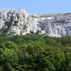 Vue du pic de Bertagne, une falaise imposante surplombant une forêt verdoyante. Un petit bâtiment est visible sur le flanc de la falaise.