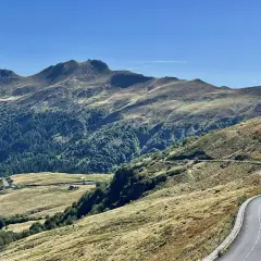 Route sinueuse dans les montagnes, avec des pâturages dorés et une forêt dense. Pas de Peyrol, un paysage grandiose sous un ciel bleu.