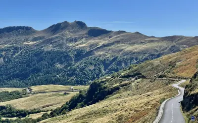 Route sinueuse dans les montagnes, avec des pâturages dorés et une forêt dense. Pas de Peyrol, un paysage grandiose sous un ciel bleu.