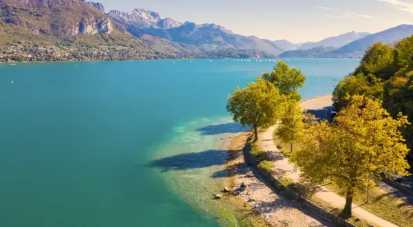 Vue aérienne du lac d'Annecy, ses eaux turquoise bordées par une route sinueuse et des arbres aux couleurs d'automne. La taille du lac est impressionnante.