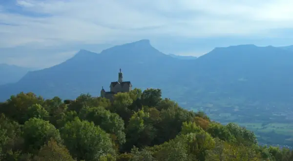 Un petit Mont Saint Michel savoyard, une chapelle perchée sur une colline boisée, domine un paysage de montagnes bleutées sous un ciel nuageux.