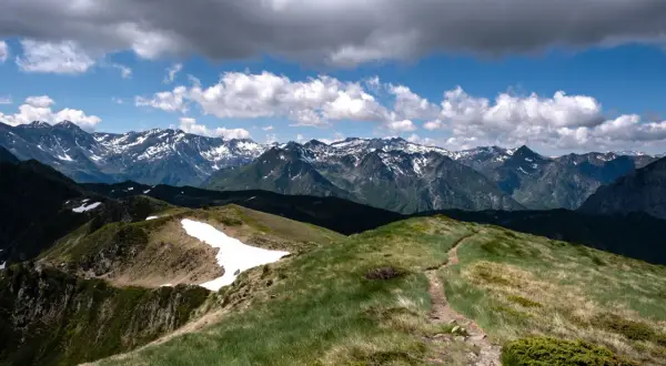 Sentier rocailleux menant vers le pic de Tarbesou, avec des montagnes enneigées en arrière-plan sous un ciel nuageux.