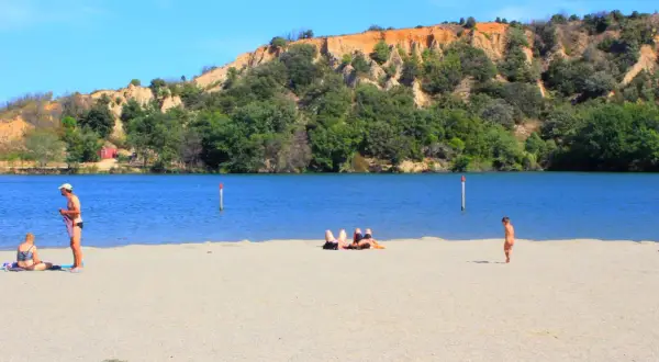 Plage de sable fin au lac de Vinça, avec des baigneurs profitant du soleil et de la vue sur la colline verdoyante.
