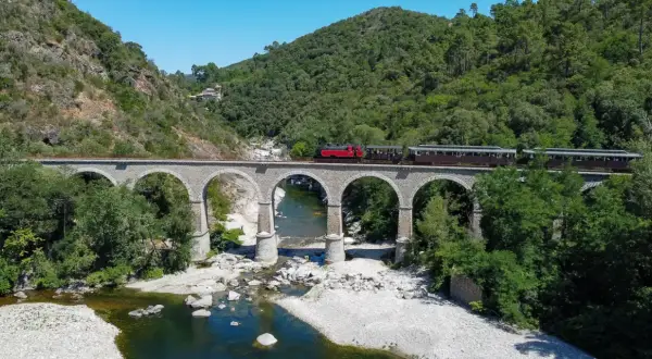 Le petit train des Cévennes traverse un viaduc de pierre au-dessus d'une rivière. La durée du trajet est une invitation à la découverte de ce paysage verdoyant.