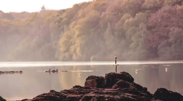 Épée plantée dans un rocher au bord du lac de Trémelin, entouré de brume et de collines boisées.