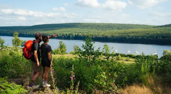 Couple randonneurs admirant le lac des Vieilles Forges, avec des voiliers sur l'eau et une forêt dense en arrière-plan.