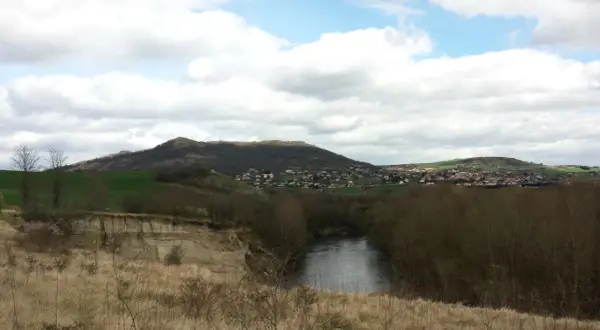 Vue panoramique d'une rivière sinueuse bordée d'arbres, avec une falaise de malmouche en contrebas et un village niché sur une colline verdoyante.