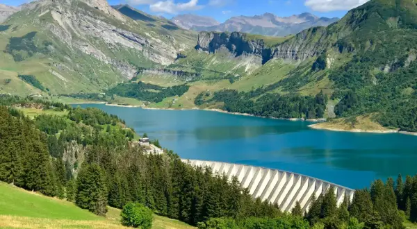Vue panoramique du lac de Roselend, un barrage imposant bordé de forêts verdoyantes et de montagnes majestueuses sous un ciel bleu éclatant.