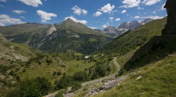 Vue panoramique d'un col de tentes dans les Pyrénées, avec une route sinueuse traversant des prairies verdoyantes et des montagnes majestueuses sous un ciel bleu parsemé de nuages.