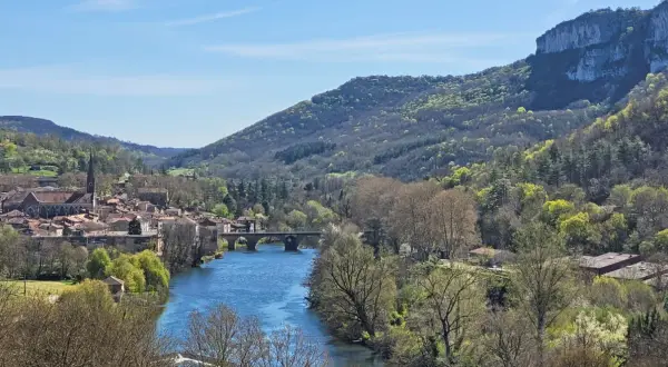 Vue panoramique de la gorge de l'Aveyron avec un village pittoresque, une rivière bleue et des collines verdoyantes sous un ciel clair.