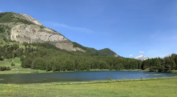 Le lac de Barbeyroux, paisible, reflète le ciel bleu. Des montagnes rocheuses et une forêt dense bordent ses rives verdoyantes.