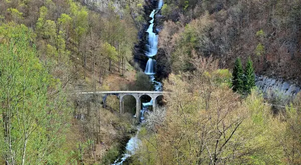 La cascade du Bief de la Ruine dévale une falaise rocheuse, traversée par un pont en arc. La nature verdoyante environnante annonce le printemps.