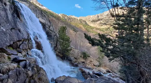 La cascade des Molières dévale une paroi rocheuse, entourée de pins et de montagnes enneigées sous un ciel bleu.