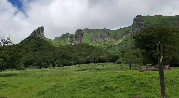 Paysage montagneux verdoyant avec des formations rocheuses spectaculaires. Un panneau indique "Fond de vallée", peut-être le chemin vers la cascade de la biche.