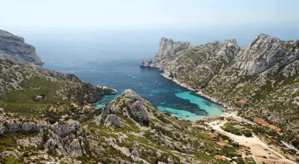 Vue aérienne de la calanque de Sormiou, un joyau bleu turquoise niché entre des falaises rocheuses. L'itinéraire pour y accéder est visible, serpentant à travers la végétation méditerranéenne.