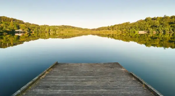 Un ponton en bois mène au lac Marion de Biarritz, reflétant les collines verdoyantes sous un ciel clair.