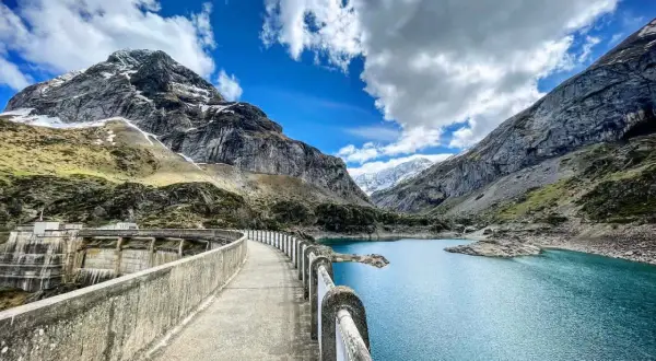 Vue panoramique du lac des gloriettes, un barrage imposant et des montagnes majestueuses sous un ciel bleu et nuageux.