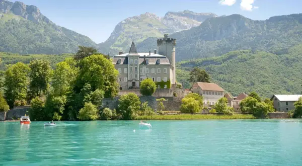 Un château majestueux surplombe un lac turquoise, invitant à aller à Annecy. Des montagnes verdoyantes en arrière-plan.