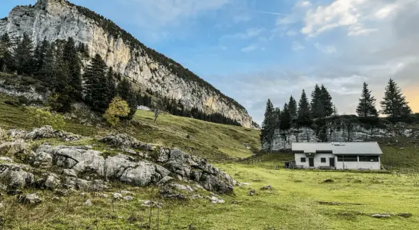 Un chalet blanc se dresse dans un pré verdoyant au pied d'une falaise imposante. Le col de l'Alpette offre un paysage alpin paisible.